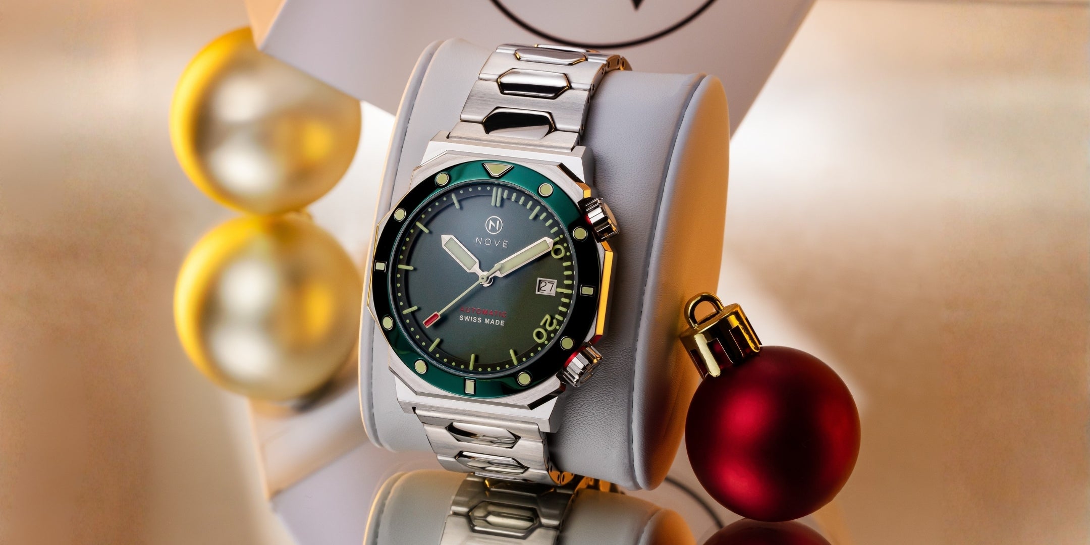 Silver watch with a green dial and bezel on a white stand with Christmas ornaments.