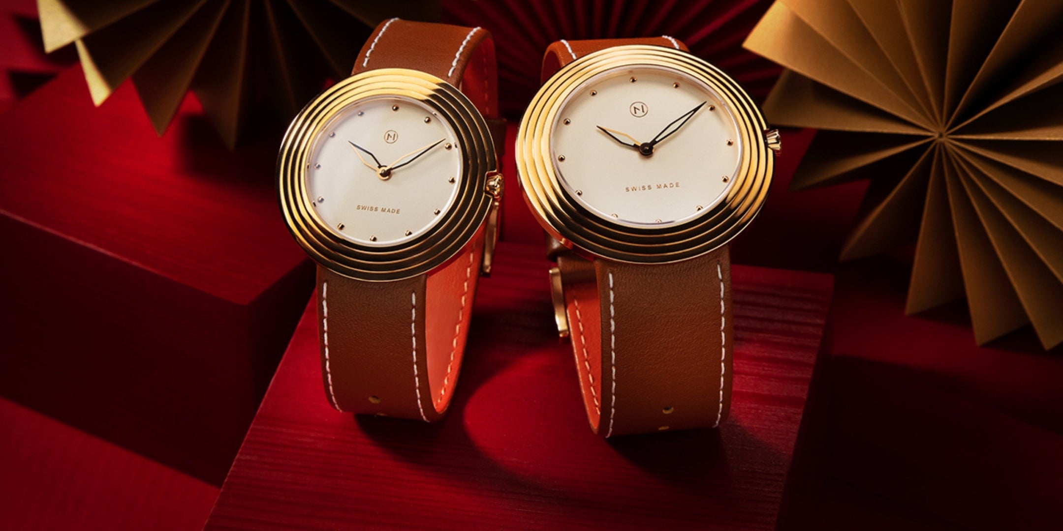 Two gold watches with brown leather straps on a red surface with decorative elements.